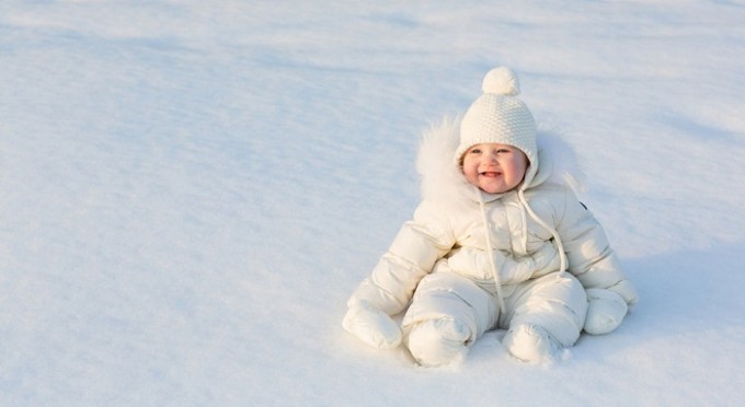 Un neonato seduto a giocare sulla neve. Il bambino indossa una tutina tutta bianca, ideale per la neve La prima neve con il neonato: consigli e precauzioni